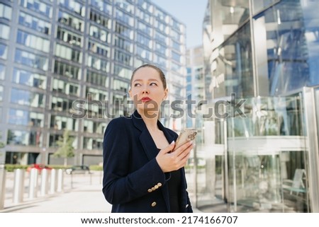 A close photo of a female realtor who is using a cell phone while waiting. A woman between the office buildings in the financial district. A businesswoman in a blazer in the cluster of tall buildings.