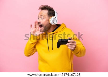 Middle age man playing with a video game controller isolated on pink background shouting with mouth wide open to the side