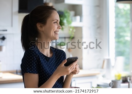 Excited happy smartphone user woman holding mobile phone, using gadget in kitchen, consulting online recipe on Internet, looking at window away, laughing, smiling, having fun