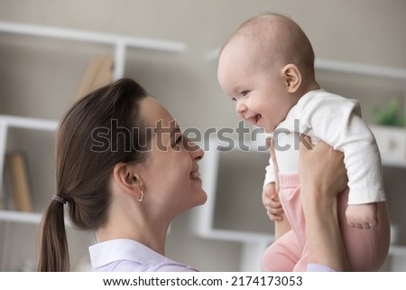 Young 25s cheerful loving mother lifts her adorable smiling three-months-old baby enjoy playtime together at home, close up shot, side profile view. Family bond, happy motherhood, childcare concept Royalty-Free Stock Photo #2174173053