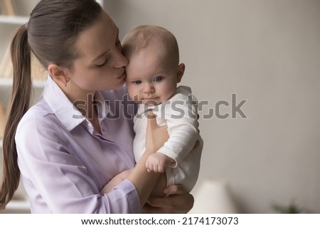 Young 25s beautiful loving mommy holding in arms, kissing her adorable baby in bodysuit, express caress, feeling love, close up shot. Happy Mothers Day, motherhood, carefree babyhood, custody concept Royalty-Free Stock Photo #2174173073