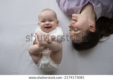 Adorable healthy three-month-old baby in bodysuit and dry diaper lying on bed near loving mother, close up top view. Carefree family resting at home on white bedsheets, feeling happy, babyhood concept Royalty-Free Stock Photo #2174173085