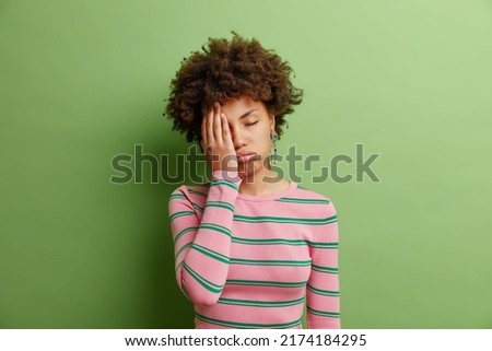 Bored young woman listens something uninteresting feels apathy has indiffernt look keeps eyes closed covers half of face with palm dressed in casual striped jumper isolated over green background
