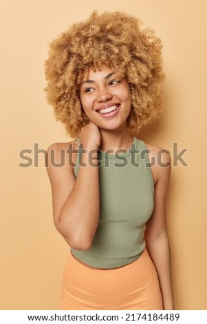 Vertical shot of happy sporty woman keeps hand on neck dressed in t shirt and leggings has good figure as goes in for sport regularly looks away smiles happily isolated over beige background Royalty-Free Stock Photo #2174184489