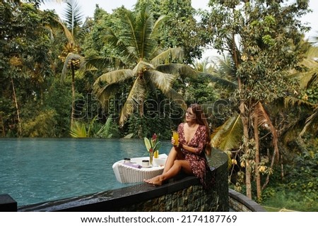 Female with long hair in red dress drink orange juice cup by swimming pool in tropical hotel. Young beautiful woman eat breakfast food in pool on private villa at luxury resort. Royalty-Free Stock Photo #2174187769