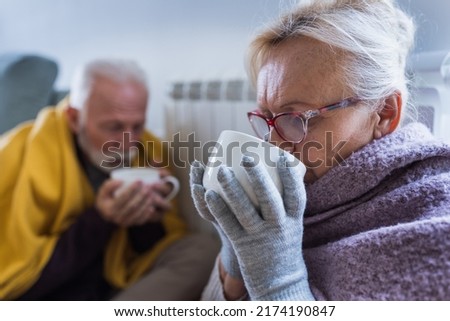 Senior couple covered with blanket sitting beside heater in living room, drinking hot tea from big cup and trying to warm in winter season and energy crisis Royalty-Free Stock Photo #2174190847