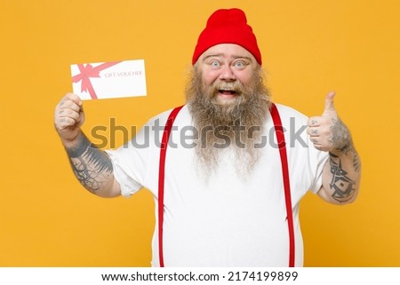 Fat pudge obese chubby overweight tattooed bearded man 30s has big belly in white t-shirt red hat suspenders hold gift voucher flyer mock up show thumb up gesture isolated on yellow background studio