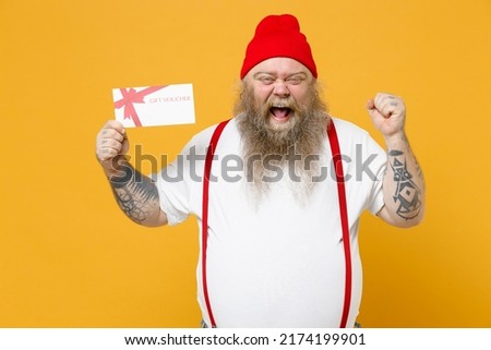 Fat pudge obese chubby overweight tattooed bearded man 30s has big belly in white t-shirt red hat hold gift voucher flyer mock up do winner gesture clench fist isolated on yellow background studio