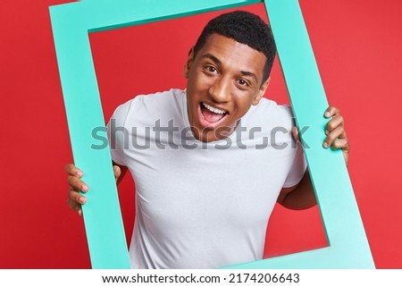 Playful mixed race man looking through a picture frame while standing against red background