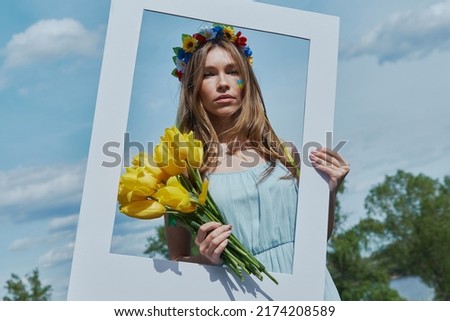 Beautiful young Ukrainian woman in floral crown holding picture frame with blue sky in the background