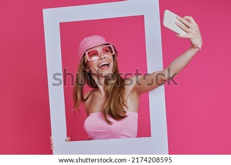 Playful young woman making selfie and looking through a picture frame while standing against pink background