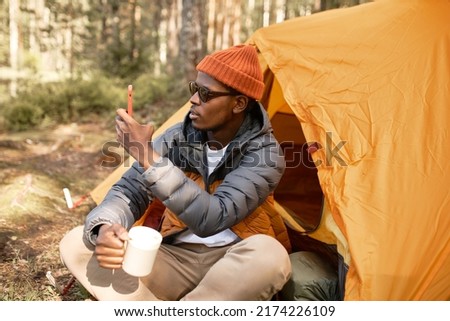 Handsome african american tourist guy in red hat and sunglasses holding mug with hot beverage and phone, taking picture sitting next to orange tent in camping. Traveling, adventures and technology