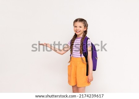 A little girl with a satchel behind her back holds your advertisement on a white isolated background on her hand. Royalty-Free Stock Photo #2174239617