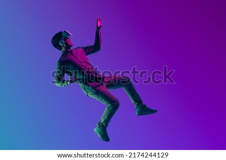 Young Asian man wearing VR headset enjoy playing video game and levitating in the air on futuristic purple cyberpunk neon light banner background. Metaverse technology concept.