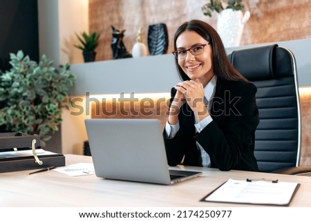 Portrait of a gorgeous smart successful caucasian brunette business woman with glasses, company ceo, top manager, sitting at a desk in a modern office, looking at the camera, smiling positively Royalty-Free Stock Photo #2174250977