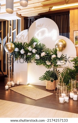 festive arch with white cake with berries, gold air balloons, white flowers, candles, monstera leaves on birthday party. photo zone in restaurant