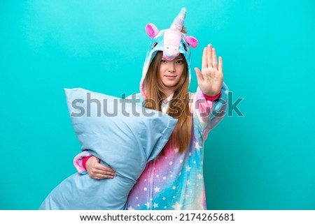 Young caucasian girl with unicorn pajamas holding pillow isolated on blue background making stop gesture