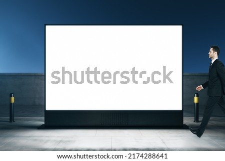 Street advertising concept with young man walking by blank white illuminated billboard with space for your logo or text on night city background, mock up