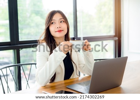 Asian young business woman enjoying coffee during in morning and smiling out the window makes me feel relaxed in my spare time from work, lunch in a modern cafe using laptop working sitting in office. Royalty-Free Stock Photo #2174302269