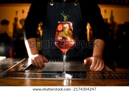 beautiful view of a glass with cold alcoholic drink with strawberries and mint decorated with branch of rosemary on bar. Bartender on background