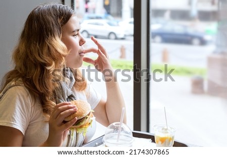 Asian young girl sitting at table indoors, holding hamburger in her hand, looking out window and from pleasure licking her fingers. Concept: fatty, high-calorie, unhealthy food Royalty-Free Stock Photo #2174307865