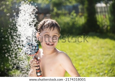 Happy kid playing with garden hose and having fun with spray of water in sunny backyard. Summer time. Kid Boy helps water garden with hose. Slow life. Enjoying the little things. Summer holiday