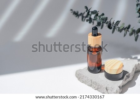 Amber bottle with dropper pipette and and jar with cream, serum or essential oil on grey concrete podium with eucalyptus branches. White background with daylight. Beauty concept for face and body care