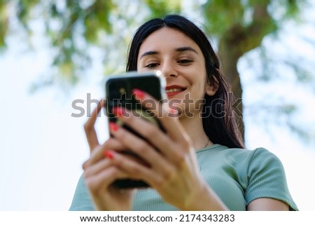Happy woman wearing tee standing on city park, outdoors looking at the phone screen and using phone. Messaging with friends, watching video or scrolling on social media. Bottom up view.
