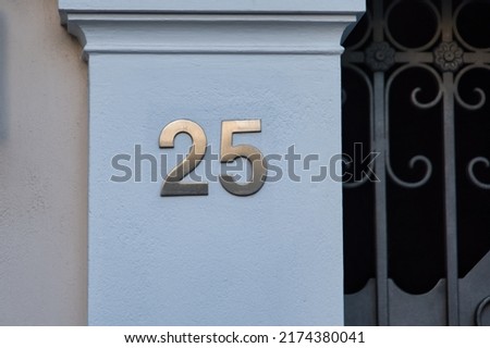 The number '25' is mounted in big golden letters on a white column next to a steel gate.  Luxurious house exterior. 