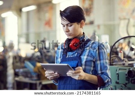 Serious busy female factory engineer with short black hair standing in workshop and using tablet for lathe examination