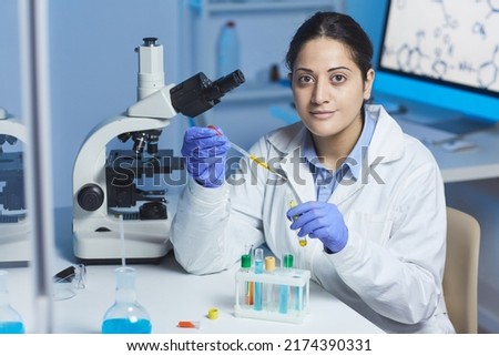 Portrait of content young Indian female pharmacist in lab coat and surgical gloves sitting at desk and mixing reagents in test tube using pipette Royalty-Free Stock Photo #2174390331