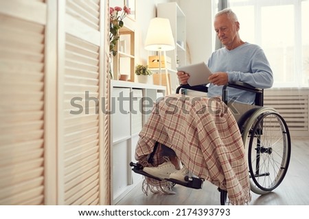 Serious concentrated disabled senior man with paralyzed legs sitting in wheelchair and reading online article on tablet