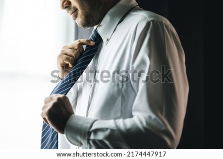 Confident businessman tying or adjust the necktie near window in hotel room in the morning. Handsome man wearing a nice necktie on wedding day. Royalty-Free Stock Photo #2174447917