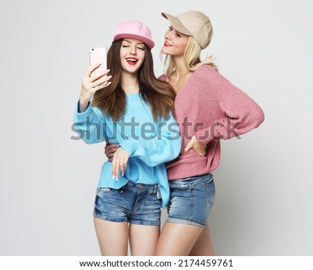 Portrait of two female friends dressed in sweaters and caps standing together and taking a selfie over grey background