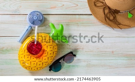 Closeup studio top view shot colorful pineapple shape rubber ring floating holder with iced cold drink glass with straw, sunglasses, small fan and weaving rattan hat with string hatband on wood table. Royalty-Free Stock Photo #2174463905