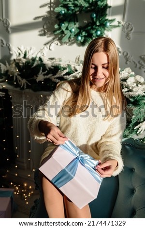A beautiful young woman with blond hair in light clothes sits on the sofa and holds a wrapped gift with a bow in her hands. Time to open Christmas presents. New Years is soon
