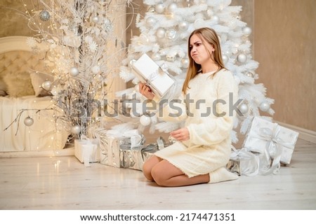 A beautiful blonde girl in a light cozy sweater opens Christmas gifts. She is in a bright bedroom decorated with a Christmas tree for Christmas.