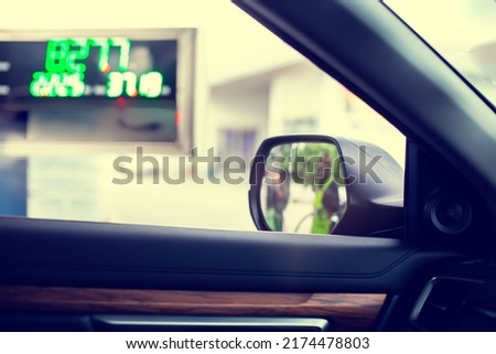 Selective focus to wing mirror with blurry fuel price display board and the officers are refueling in gas station.