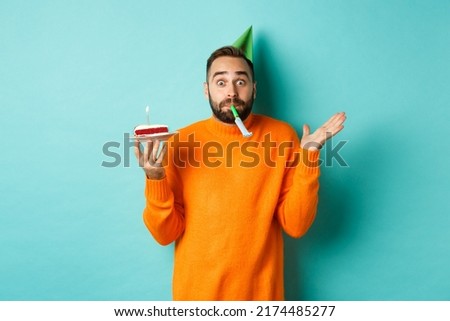Happy birthday guy celebrating, wearing party hat, blowing wistle and holding bday cake, standing against white background