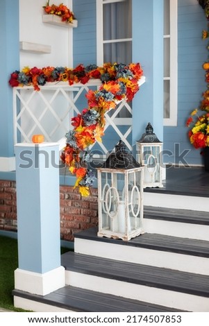 House entrance staircase decorated for autumn holidays, fall flowers and pumpkins. Cozy porch of the house with wooden lanterns in fall time. Halloween design home with yellow fall leaves and lamps