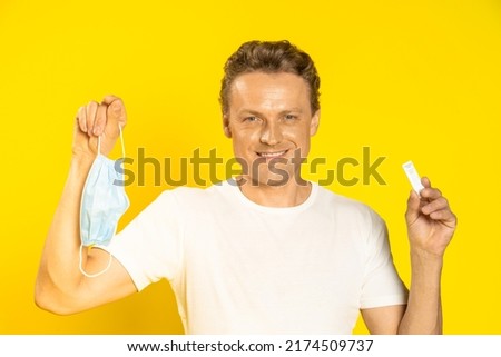 Happy man holding test and medical face mask in hands celebrating victory pandemic coronavirus or monkeypox. Handsome man in white t-shirt and medical mask on yellow background. Healthcare concept. 