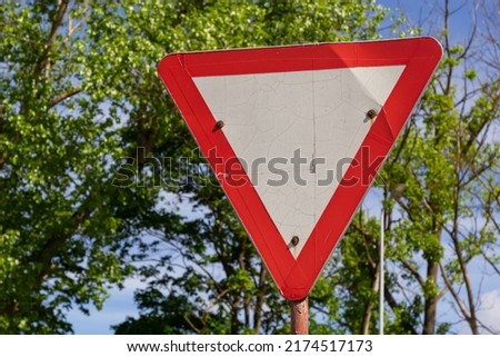 Close-up of give way road sign. Red and white triangle. Green trees and blue sky on background.