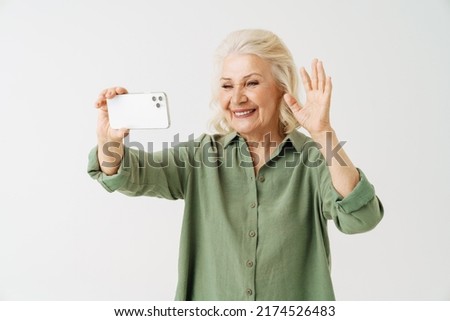 Grey senior woman gesturing while taking selfie on mobile phone isolated over white background