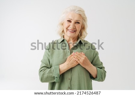 Grey senior woman in shirt smiling while holding her hands on chest isolated over white background Royalty-Free Stock Photo #2174526487