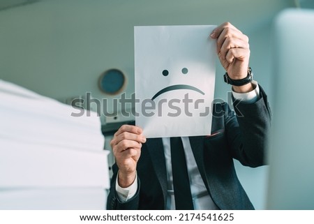 Unhappy businessman holding paper with frowning emoticon in office interior, selective focus Royalty-Free Stock Photo #2174541625