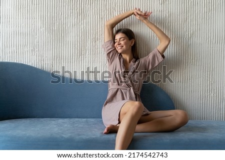Indoor portrait of relaxed woman in dressing gown having rest on blue couch stretching hands up, smiling with closed eyes on sunday morning with copy space on the right side of picture Royalty-Free Stock Photo #2174542743