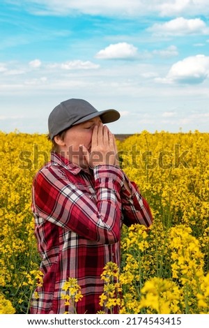 Female farmer with pollen allergy sneezing in blooming rapeseed field. Farm worker wearing plaid shirt and trucker's hat with hands covering nose. Selective focus. Royalty-Free Stock Photo #2174543415