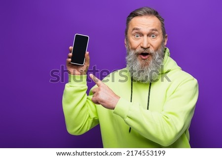 amazed senior man with beard and grey hair pointing at smartphone isolated on purple