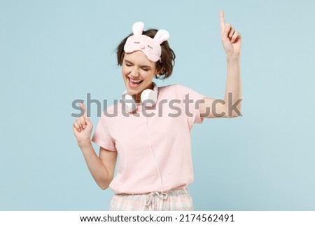 Young caucasian woman 20s in pajamas jam sleep eye mask headphones rest relaxing at home listen to music dancing spread hands isolated on pastel blue background studio. Good mood night bedtime concept