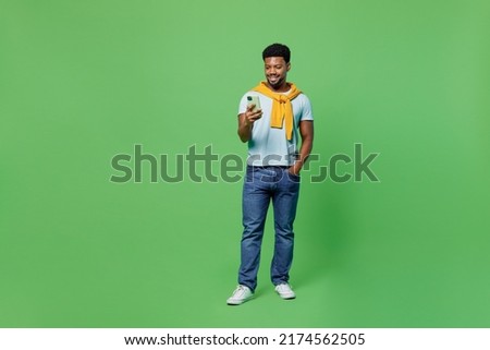 Full body smiling happy fun young man of African American ethnicity 20s in blue t-shirt hold in hand use mobile cell phone isolated on plain green background studio portrait. People lifestyle concept